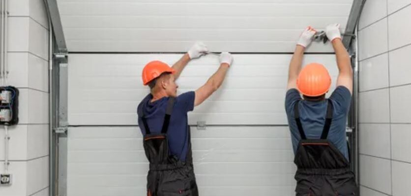 Two men in overalls and hard hats are collaboratively repairing a garage door.