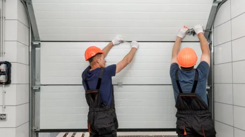 Two men in overalls and hard hats are collaboratively repairing a garage door.