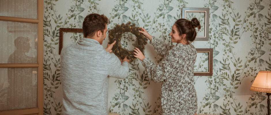 A man and woman are decorating a wall with frames and a brown wreath. The wall has wallpaper with flowery pattern.